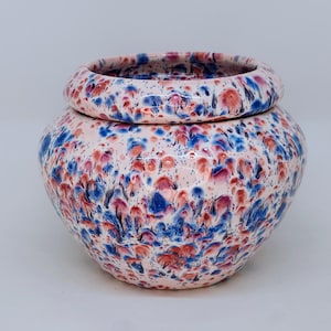 May include: A white ceramic planter with a speckled design in shades of blue, pink, and red. The planter has a wide, round base and a slightly narrower opening.