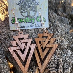 May include: A pair of wooden earrings with a geometric triangle design. The earrings are made of real wood and have silver hooks. The earrings are made by Sawmill Naturals Jewelry Co. and are made in the USA.