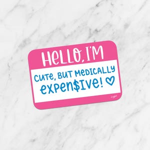 May include: A pink name tag sticker with the text "HELLO, I'M" in white, and "cute, BUT MEDICALLY expen$ive!" in blue. A small blue heart is at the end of the text. The sticker is on a white and gray marble background.