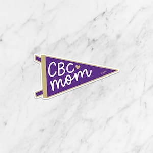 May include: Purple pennant with gold text that reads "CBC mom" with a small gold heart between the words. The pennant has a gold border and a white background.