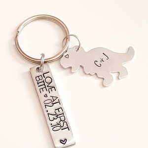May include: Silver keychain with a dinosaur charm and a rectangular tag that reads "Love at First Bite 02.23.10" with a heart symbol. The dinosaur charm has the initials "C+J" engraved on it.