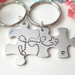 May include: A silver puzzle keychain with two pieces, one with the outline of California and the other with the outline of Texas. The pieces are connected by a dotted line with a heart at the end. The keychain has the letters "C" and "R" on each piece.