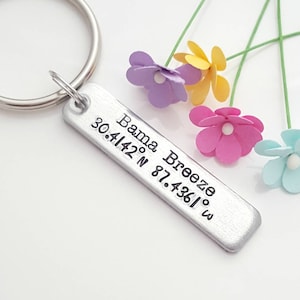 May include: Silver keychain with a personalized tag that reads "Bama Breeze 30.4142° N 87.4361° W". The keychain is surrounded by four small paper flowers in pink, yellow, purple, and blue.