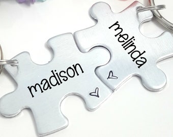 May include: Two silver puzzle pieces with the names "madison" and "melinda" engraved on them. The pieces are connected by two small hearts and attached to a silver key ring.