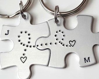 May include: Two silver puzzle piece keychains with a dotted line connecting the pieces and a heart at the end of each line. The keychains are labeled with the letters "J" and "M".