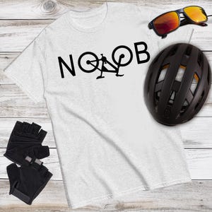May include: A light gray t-shirt with the word "NOOB" in black, featuring a bicycle graphic. The image also includes black cycling gloves, a black helmet, and sunglasses with orange lenses.