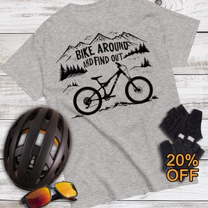 May include: A gray t-shirt with a mountain bike graphic and the text "BIKE AROUND AND FIND OUT." The image also shows a black helmet, sunglasses, and cycling gloves. The text "Mountain Bike T-Shirts" and "JustMeMTB.com" are also visible.