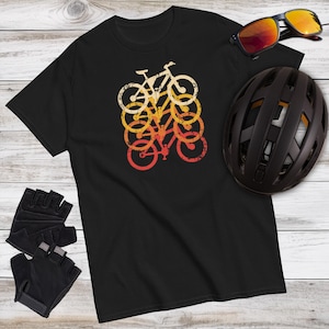 May include: Black t-shirt featuring a retro bicycle graphic in shades of orange, yellow, and white. The image also includes black cycling gloves, a black helmet, and sunglasses with orange lenses. The items are arranged on a light wood-grain surface.