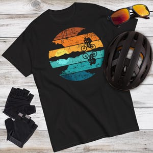 May include: Black t-shirt with a circular graphic of mountain bikers against a sunset in orange, blue, and teal. Accessories include black sunglasses, a black helmet, and black cycling gloves. The shirt is made of a soft material.
