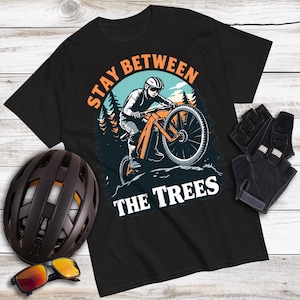 May include: Black t-shirt with a mountain biking graphic. The design features a cyclist riding an orange mountain bike with the text "STAY BETWEEN THE TREES" in orange and white. A brown helmet, sunglasses, and black gloves are also visible.
