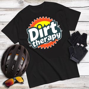 May include: Black t-shirt with the words "Dirt therapy" in a white and teal design, surrounded by an orange and yellow gear-shaped graphic. A black bike helmet, sunglasses, and black cycling gloves are also visible.