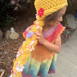 May include: A yellow crocheted hat with a long, thick braided yarn ponytail attached. The braid is decorated with various small fabric flowers in shades of pink, purple, orange, and yellow, along with thin light purple ribbon accents.