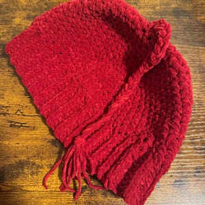 May include: A vibrant red, handmade crochet hat with a textured, plush appearance. The hat features a braided accent and a tassel detail. The hat is designed with a folded brim and is suitable for cold weather.