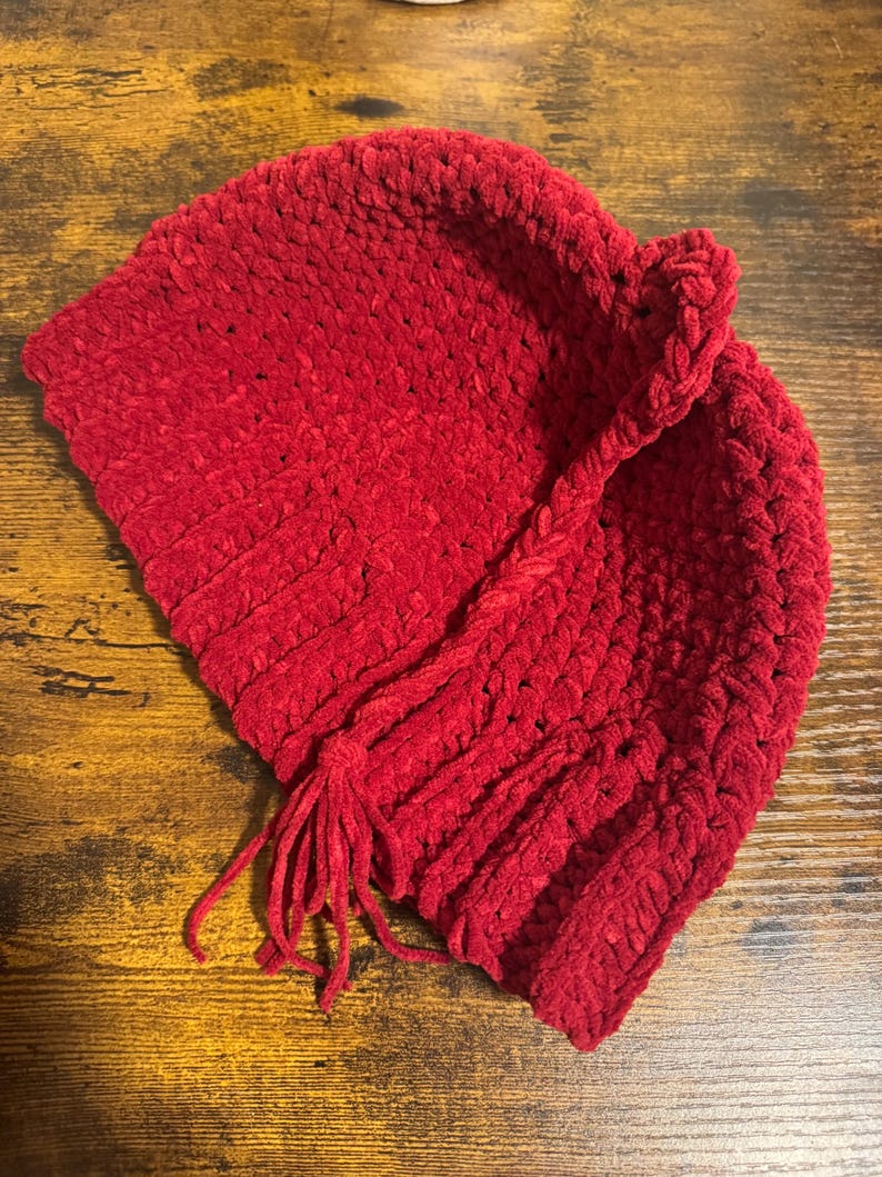 May include: A vibrant red, handmade crochet hat with a textured, plush appearance. The hat features a braided accent and a tassel detail. The hat is designed with a folded brim and is suitable for cold weather.