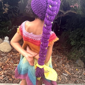 May include: A purple crocheted hat featuring a long, thick, braided ponytail adorned with small gold-colored rings. The hat is paired with a sleeveless, multi-colored crocheted dress that transitions through shades of pink, orange, yellow, and blue.
