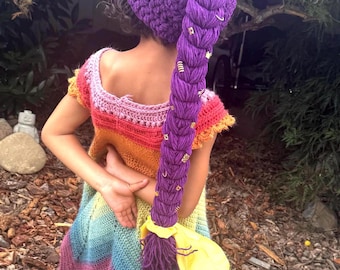 May include: A purple crocheted hat featuring a long, thick, braided ponytail adorned with small gold-colored rings. The hat is paired with a sleeveless, multi-colored crocheted dress that transitions through shades of pink, orange, yellow, and blue.