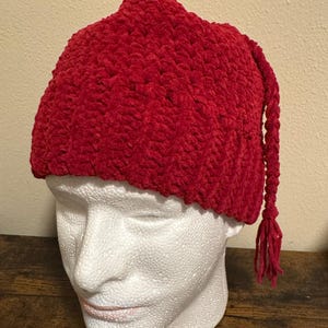 May include: A vibrant red knit hat with a textured surface and a long, braided tassel. The hat is displayed on a white mannequin head. The hat's design is a classic style, suitable for cold weather.