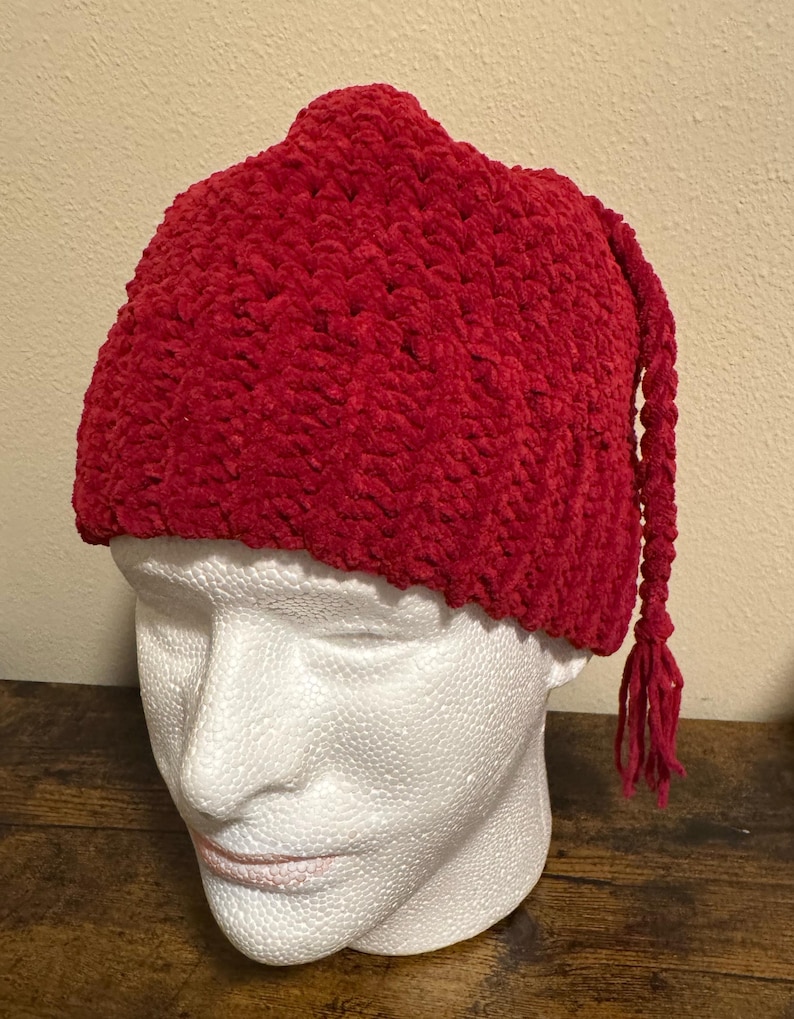 May include: A vibrant red knit hat with a textured surface and a long, braided tassel. The hat is displayed on a white mannequin head. The hat's design is a classic style, suitable for cold weather.
