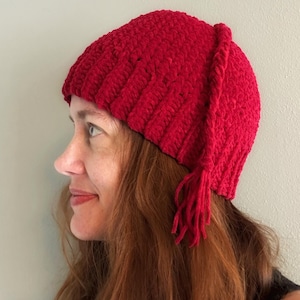 Puede incluir: Un gorro rojo vibrante, hecho a mano con ganchillo, con un diseño acanalado texturizado. El gorro presenta un pequeño detalle de borla en el lateral. Un accesorio de invierno.