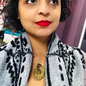 May include: A woman wearing a gray and black patterned sweater with a gold chain necklace featuring a large, fossilized ammonite pendant.