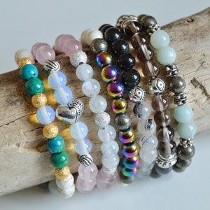 May include: A stack of eight beaded bracelets in various colors, including turquoise, pink, white, black, brown, and green. The bracelets are made with different types of beads, including gemstone beads, metal beads, and glass beads.