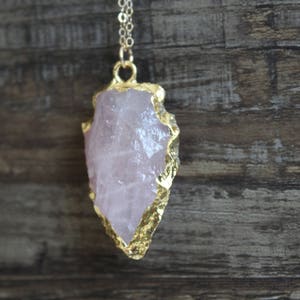 May include: A gold-plated pendant featuring a large, raw rose quartz crystal. The crystal is a light pink color and has a natural, irregular shape.