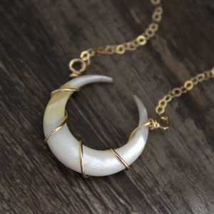 May include: A crescent moon-shaped pendant made of iridescent white shell, wrapped in thin gold-colored wire. The pendant is attached to a delicate gold-colored metal chain, featuring a smooth, polished surface with subtle cream and pearlescent tones.