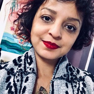 May include: A woman with curly dark hair and red highlights is wearing a gray and black patterned cardigan. She is wearing a necklace with a large, brown and gold ammonite fossil pendant.