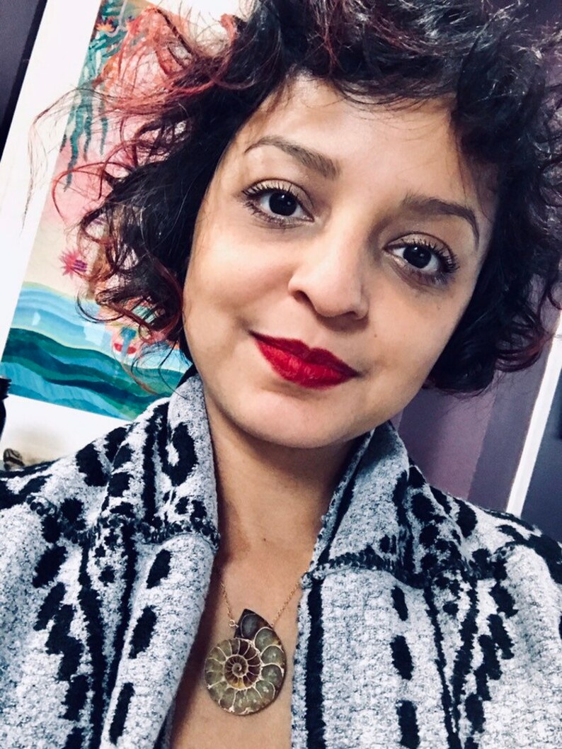 May include: A woman with curly dark hair and red highlights is wearing a gray and black patterned cardigan. She is wearing a necklace with a large, brown and gold ammonite fossil pendant.