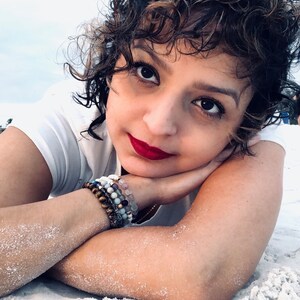 May include: A woman with short, curly brown hair is lying on her side on a white sandy beach. She is wearing a white t-shirt and has several bracelets on her wrist. Her eyes are closed and she is smiling.