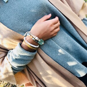 May include: A close-up of a person's wrist wearing a stack of bracelets. The bracelets are made of various materials, including beads, leather, and metal. The bracelets are a mix of colours, including white, blue, and gold.