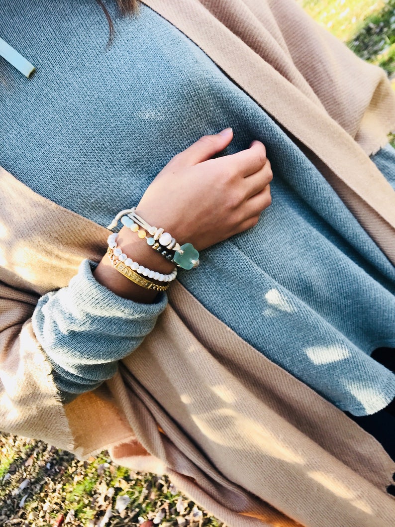 May include: A close-up of a person's wrist wearing a stack of bracelets. The bracelets are made of various materials, including beads, leather, and metal. The bracelets are a mix of colours, including white, blue, and gold.