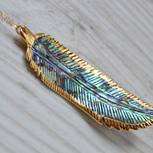 May include: A gold chain necklace with a large iridescent abalone shell feather pendant. The feather is detailed with gold accents and has a natural, organic shape.