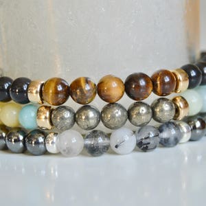 May include: A stack of four beaded bracelets featuring a variety of coloured stones, including black, brown, gold, grey and white.