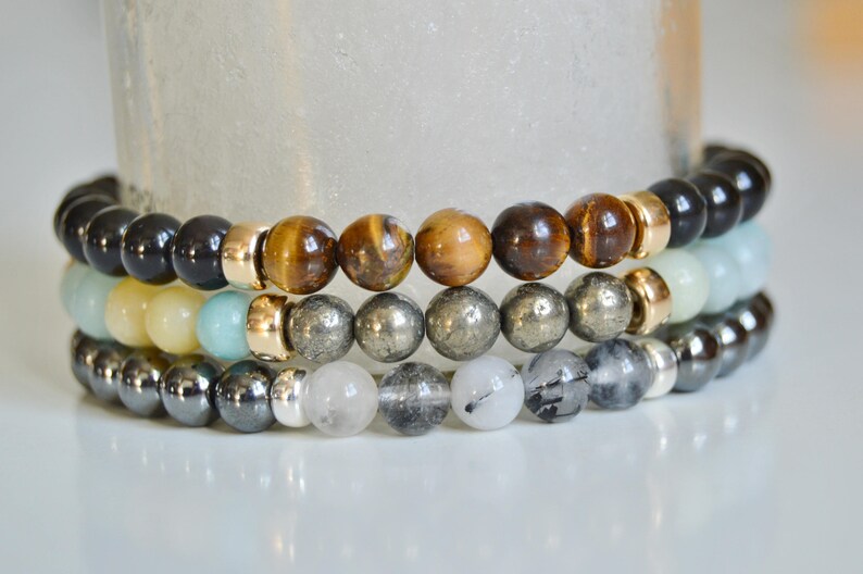May include: A stack of four beaded bracelets featuring a variety of coloured stones, including black, brown, gold, grey and white.