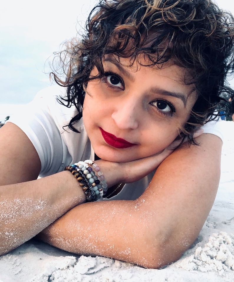 May include: A woman with short, curly brown hair is lying on her side on a white sandy beach. She is wearing a white t-shirt and has several bracelets on her wrist. Her eyes are closed and she is smiling.