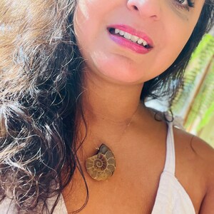 May include: A woman wearing a white top with a gold chain necklace featuring a large, brown ammonite fossil pendant.