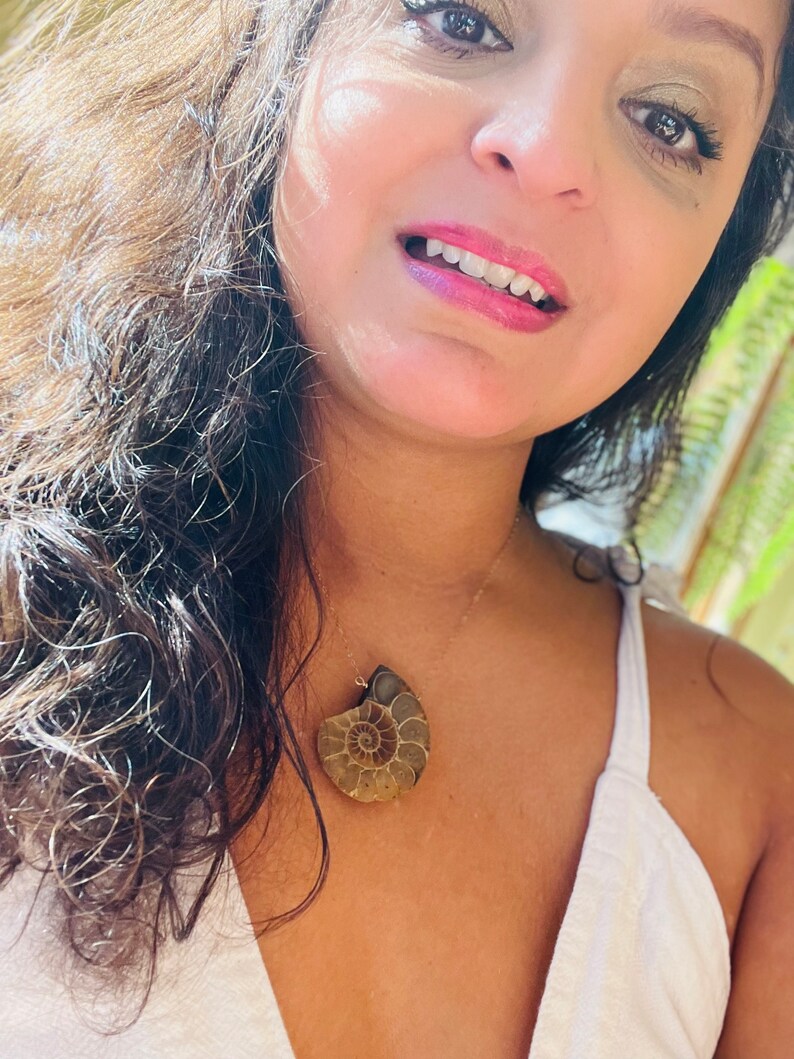 May include: A woman wearing a white top with a gold chain necklace featuring a large, brown ammonite fossil pendant.