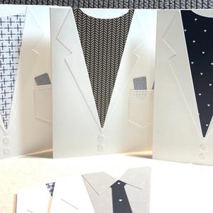 May include: White greeting cards designed to resemble suit jackets, with various patterned ties and pocket squares. Each card has a pocket and two buttons. The ties feature cross, herringbone, and heart patterns.