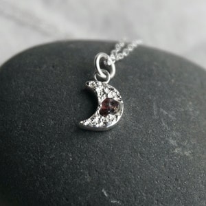 Puede incluir: Un collar de plata en forma de media luna con una pequeña piedra roja en el centro. La luna está decorada con pequeñas piedras blancas.