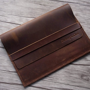 Peut inclure: Une pochette pour ordinateur portable en cuir marron avec une fermeture &agrave; rabat et une sangle cousue. La pochette porte le nom "Zachary K. Choi" en relief sur le devant.
