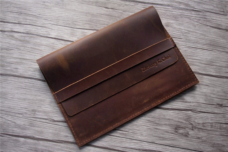 Peut inclure: Une pochette pour ordinateur portable en cuir marron avec une fermeture &agrave; rabat et une sangle cousue. La pochette porte le nom "Zachary K. Choi" en relief sur le devant.