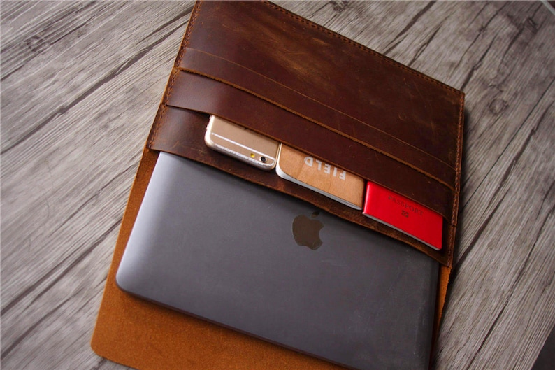 Peut inclure: Une pochette pour ordinateur portable en cuir marron avec plusieurs poches. La pochette est ouverte et un ordinateur portable, un t&eacute;l&eacute;phone et des cartes sont visibles &agrave; l'int&eacute;rieur.