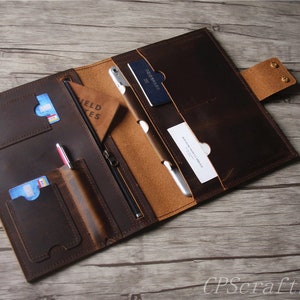 May include: A brown leather portfolio with multiple pockets and slots for cards, a pen, and a tablet. The portfolio has a zipper closure and a flap with two brass rivets.