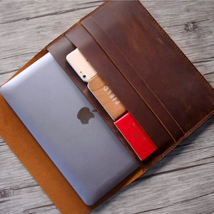 Peut inclure: Une pochette pour ordinateur portable en cuir marron avec une fermeture &agrave; rabat. La pochette a une poche sur le devant pour un t&eacute;l&eacute;phone ou d'autres petits objets. La pochette est ouverte et un ordinateur portable, un t&eacute;l&eacute;phone et un porte-cartes sont visibles &agrave; l'int&eacute;rieur.