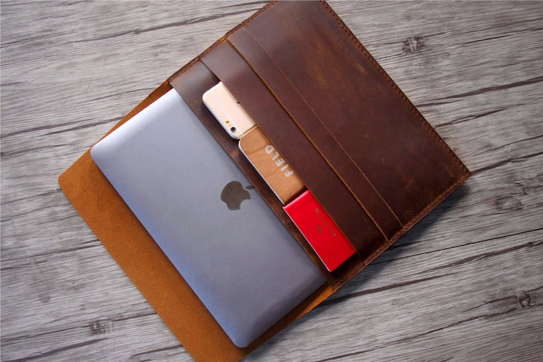 Peut inclure: Une pochette pour ordinateur portable en cuir marron avec une fermeture &agrave; rabat. La pochette a une poche sur le devant pour un t&eacute;l&eacute;phone ou d'autres petits objets. La pochette est ouverte et un ordinateur portable, un t&eacute;l&eacute;phone et un porte-cartes sont visibles &agrave; l'int&eacute;rieur.