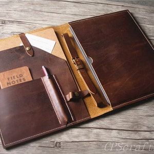 May include: A brown leather portfolio with multiple pockets and a strap. The portfolio is open and shows a notebook, pen, and a small notebook that says "Field Notes".