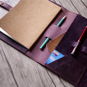 May include: A purple leather notebook cover with a brown notebook, a green pencil, a red pen, and a credit card inside. The cover has a flap that closes with a snap.