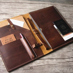 May include: A brown leather portfolio with multiple pockets and a strap closure. The portfolio is open and shows a notebook, pen, and passport inside.