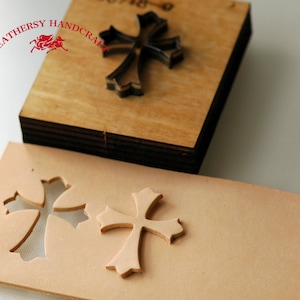 May include: A close-up of a leather craft tool with a cross-shaped design. The tool is made of wood and metal and is used to cut leather. The tool is shown next to a piece of leather that has been cut with the tool.
